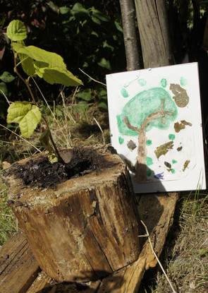 Ein gemaltes Bild mit Blättern und ein gepflanzter Baum in einer Holzscheibe. Stück von der Kinder Werkstattgruppe.,